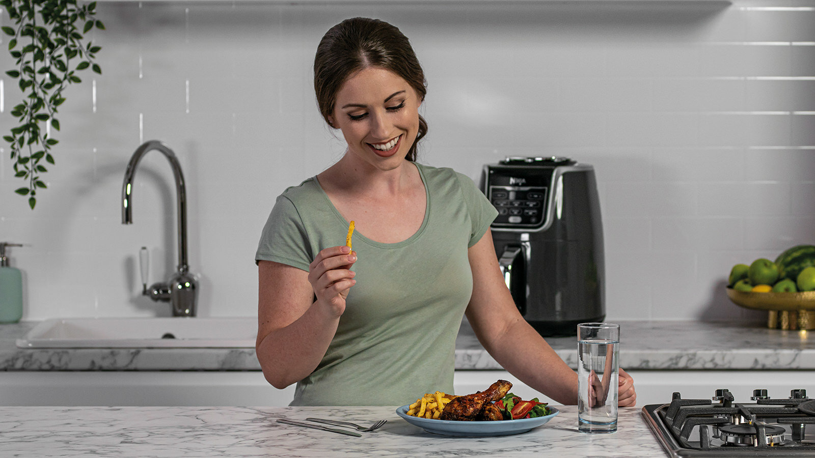 Kobieta je frytkę, tło kuchni, frytkownica, jedzenie i szklanka wody na stole.