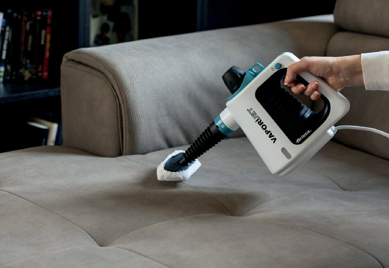 Dłoń trzymająca biało-turkusowy parowy środek czyszczący nad beżową kanapą.