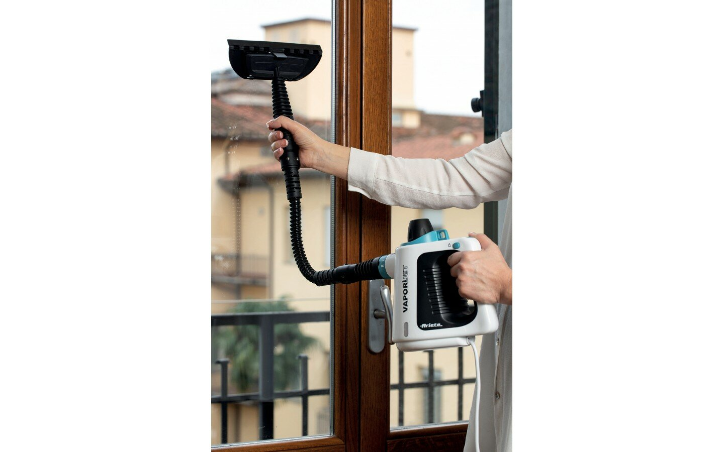 Osoba używająca biało-niebieskiego ręcznego parowego urządzenia czyszczącego na oknie. Okno ma drewnianą ramę.