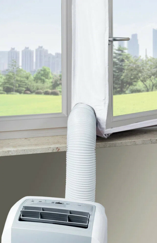 Biały klimatyzator z białym wężem wentylacyjnym z otwartego okna. Tło to zielone pole i budynki.
