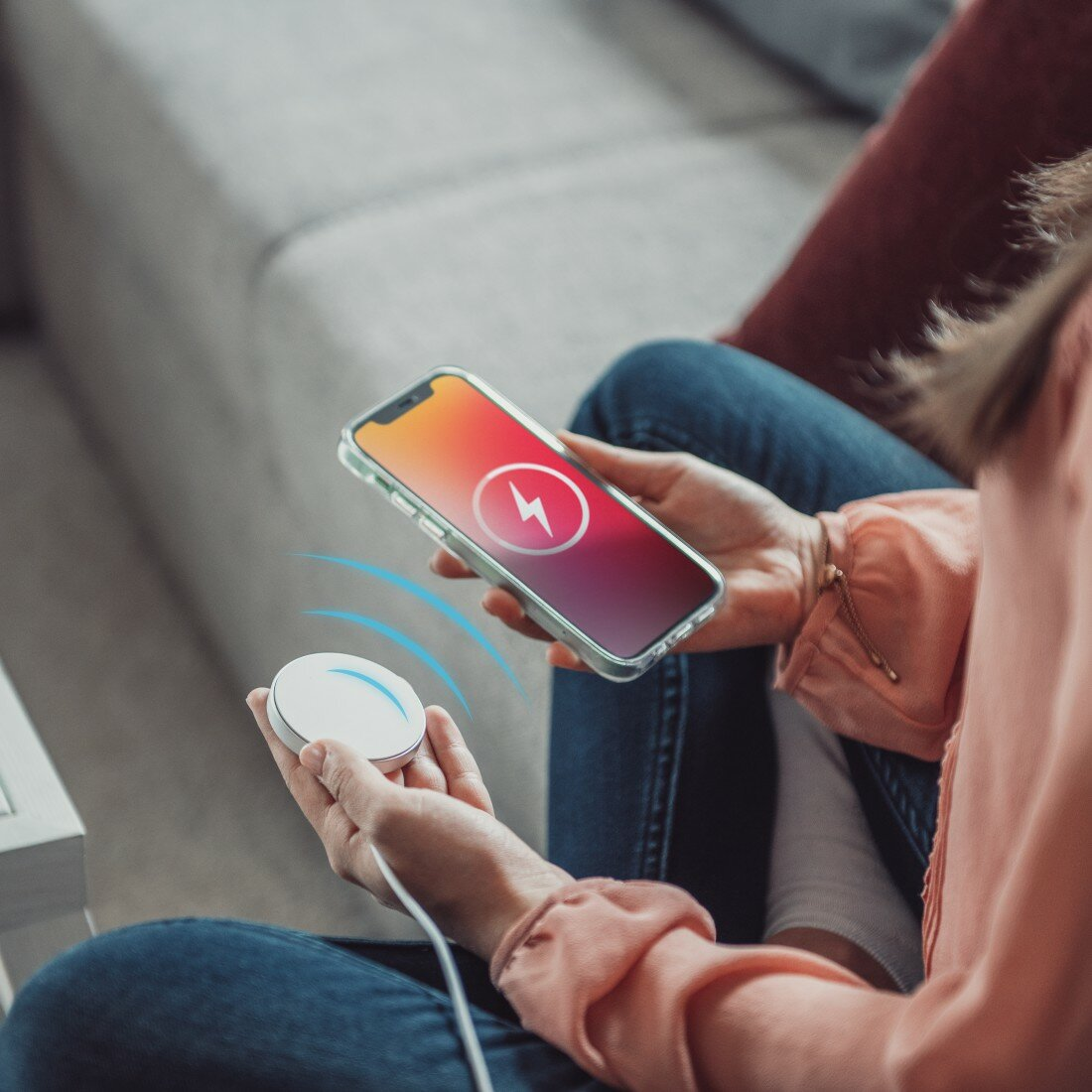 Dłonie trzymają smartfon z ikoną ładowania i białą ładowarką bezprzewodową.