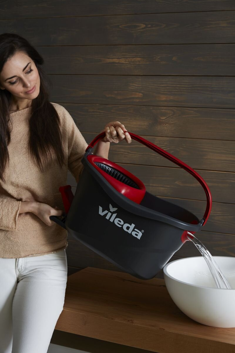 Kobieta wlewa wodę z czarno-czerwonego wiadra do białej miski. Drewniane tło.