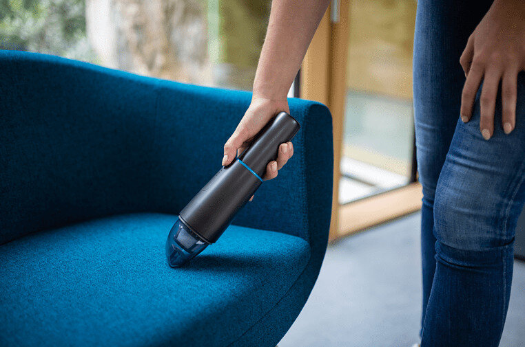 Osoba używa małego czarnego odkurzacza na niebieskim fotelu, czyszcząc kurz.