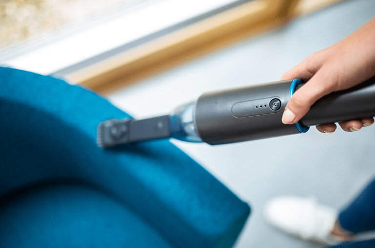 Ręka trzymająca odkurzacz, czyszcząca niebieską sofę. Tło to rozmyte okno.