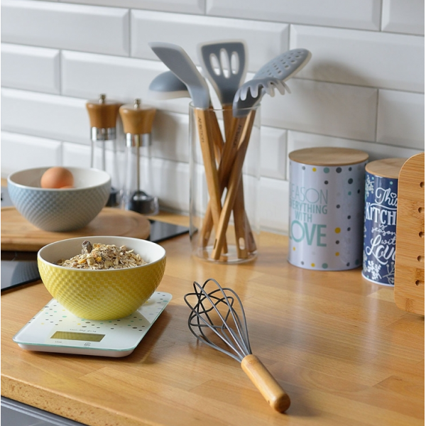 Scena kuchenna z miską owsianki, trzepaczką i innymi przyborami na drewnianej powierzchni. Białe kafelki.