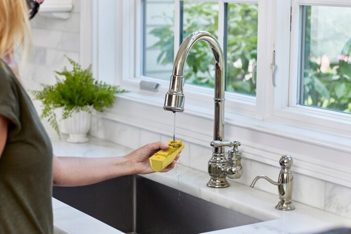 Osoba testująca wodę z kranu kuchennego żółtym urządzeniem. Biały blat i okno.