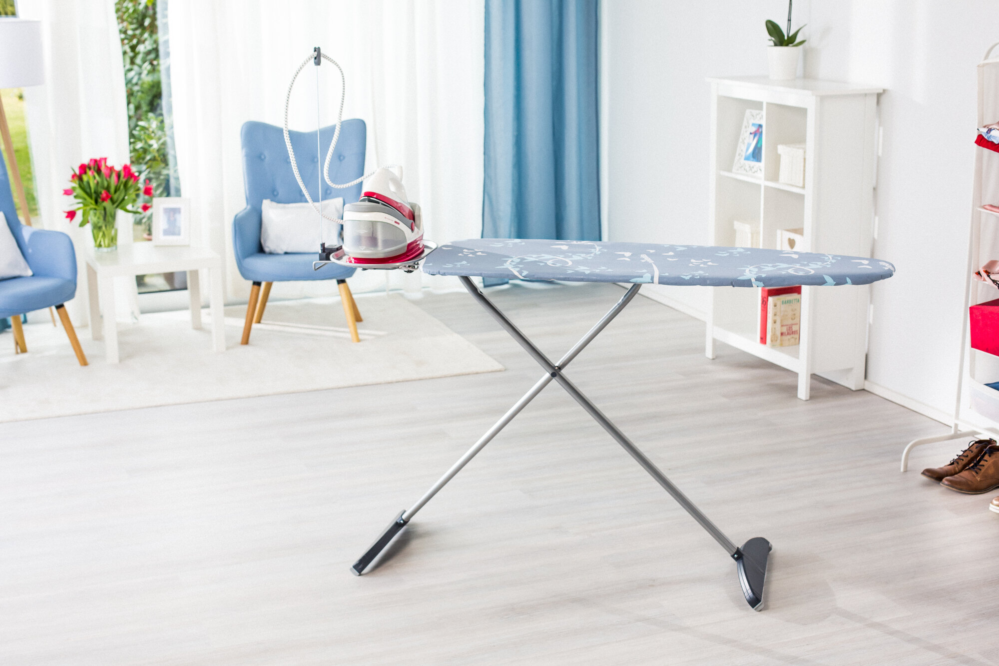 Deska do prasowania z żelazkiem parowym po lewej stronie. W tle salon z niebieskim krzesłem.