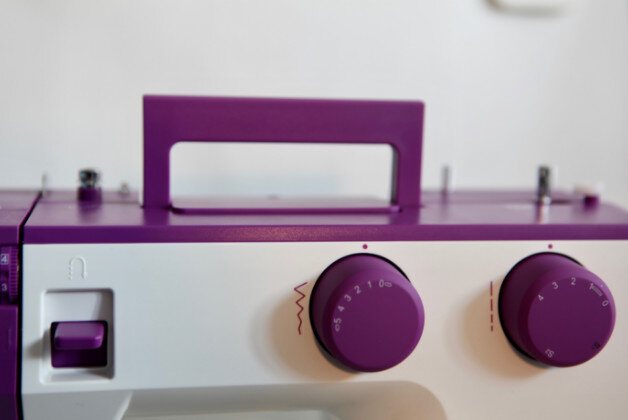 Zbliżenie maszyny do szycia. Ma fioletowy uchwyt i pokrętła. Maszyna jest biało-fioletowa.