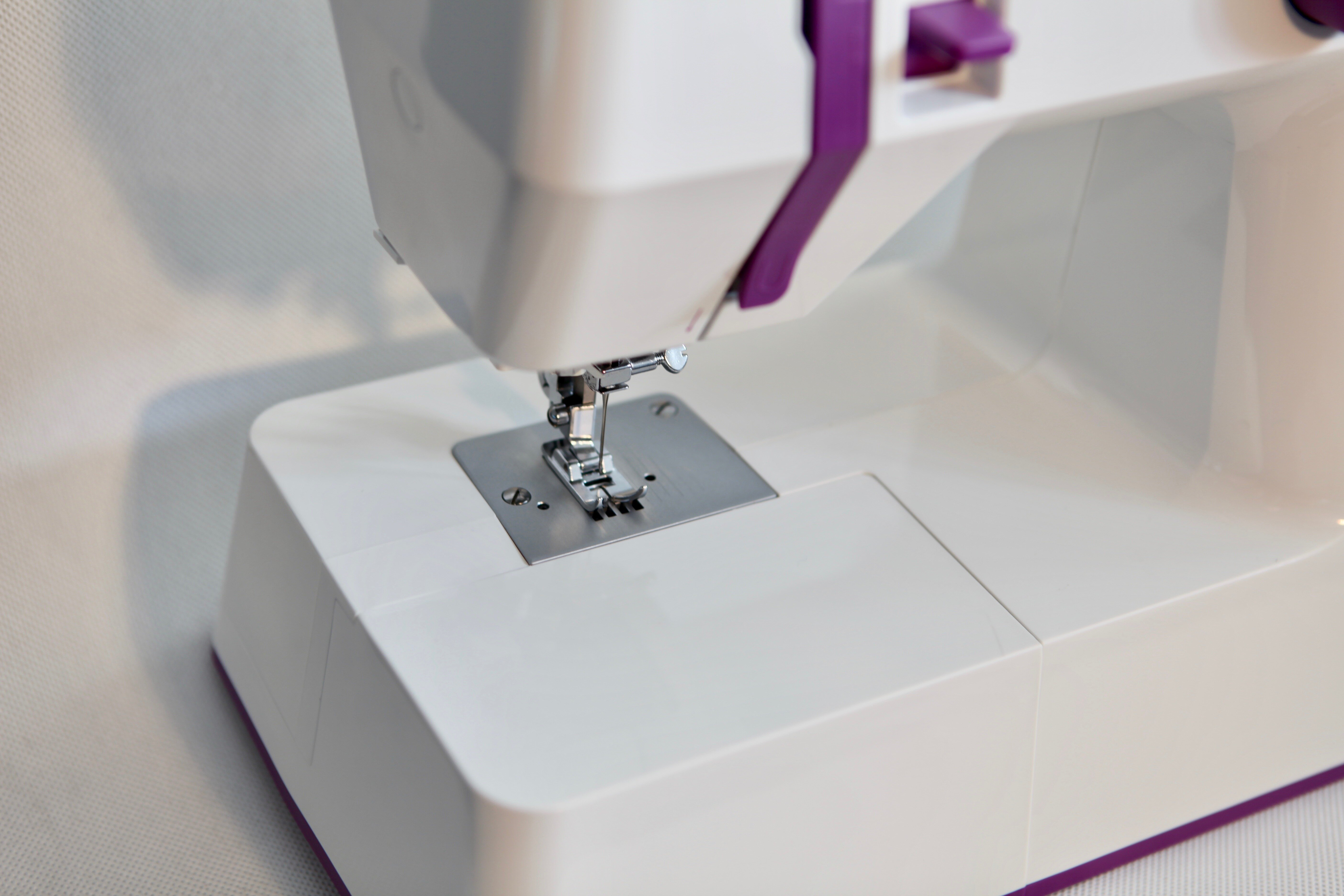Biała maszyna do szycia, z fioletowymi akcentami. Igła jest widoczna, a maszyna stoi na białej powierzchni na białym tle.