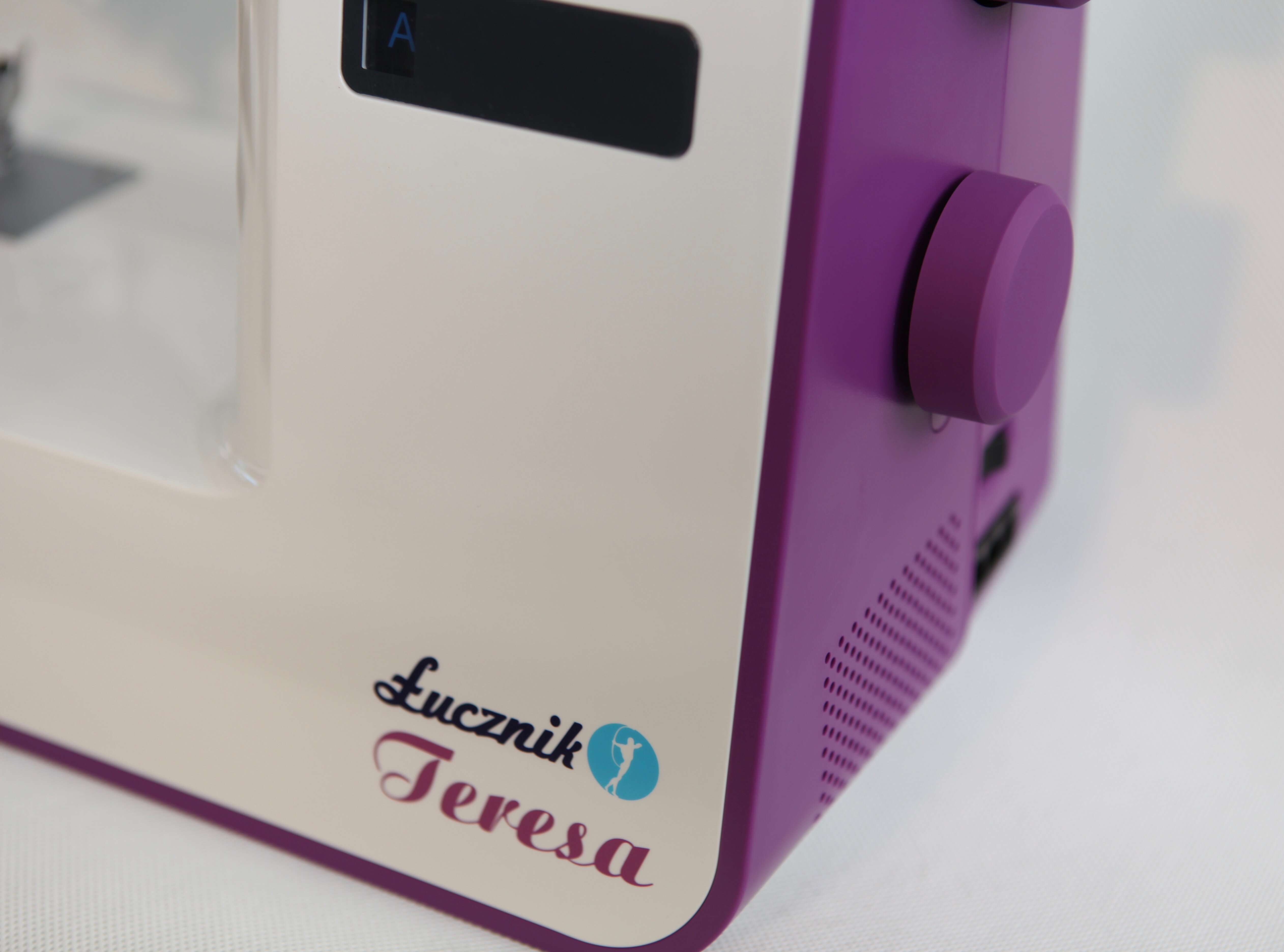 Maszyna do szycia z białą i fioletową obudową. Nazwa marki to 'Lucznik Teresa'. Z boku znajduje się pokrętło.