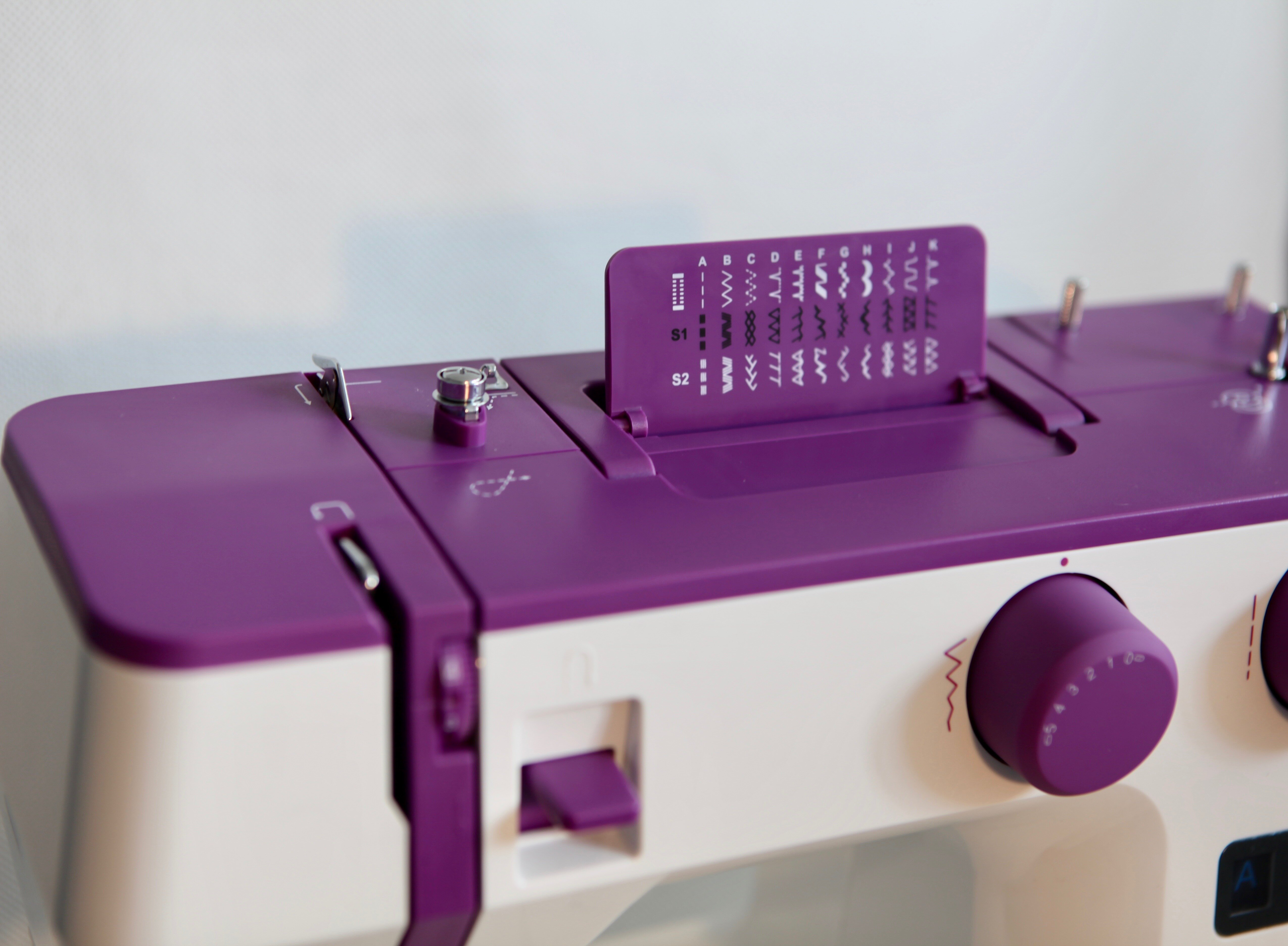 Fioletowo-biała maszyna do szycia z opcjami ściegów. Maszyna ma pokrętło i małe fioletowe drzwiczki.
