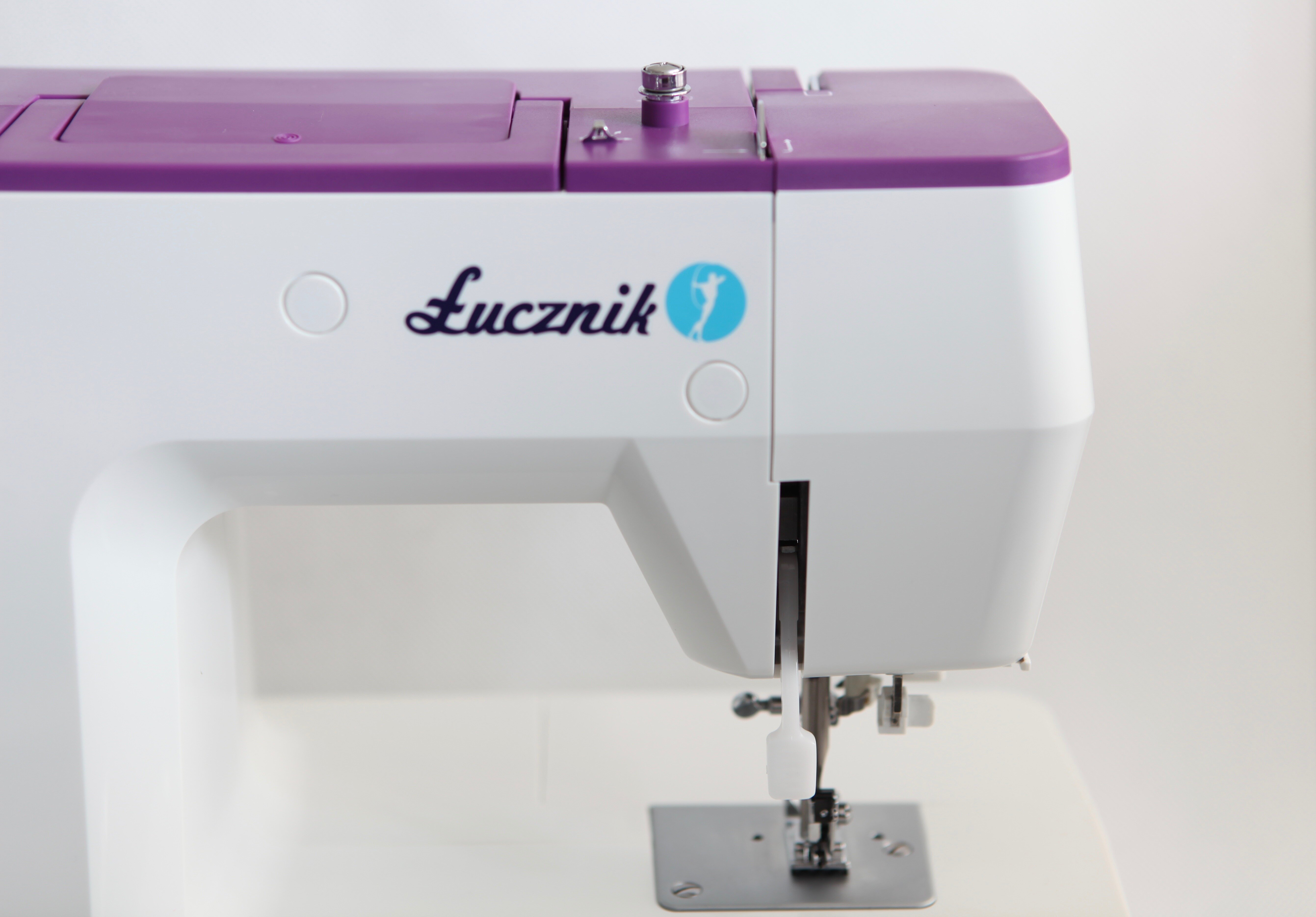 Biała i fioletowa maszyna do szycia Lucznik na białej powierzchni. Ma igłę i szpulkę nici.