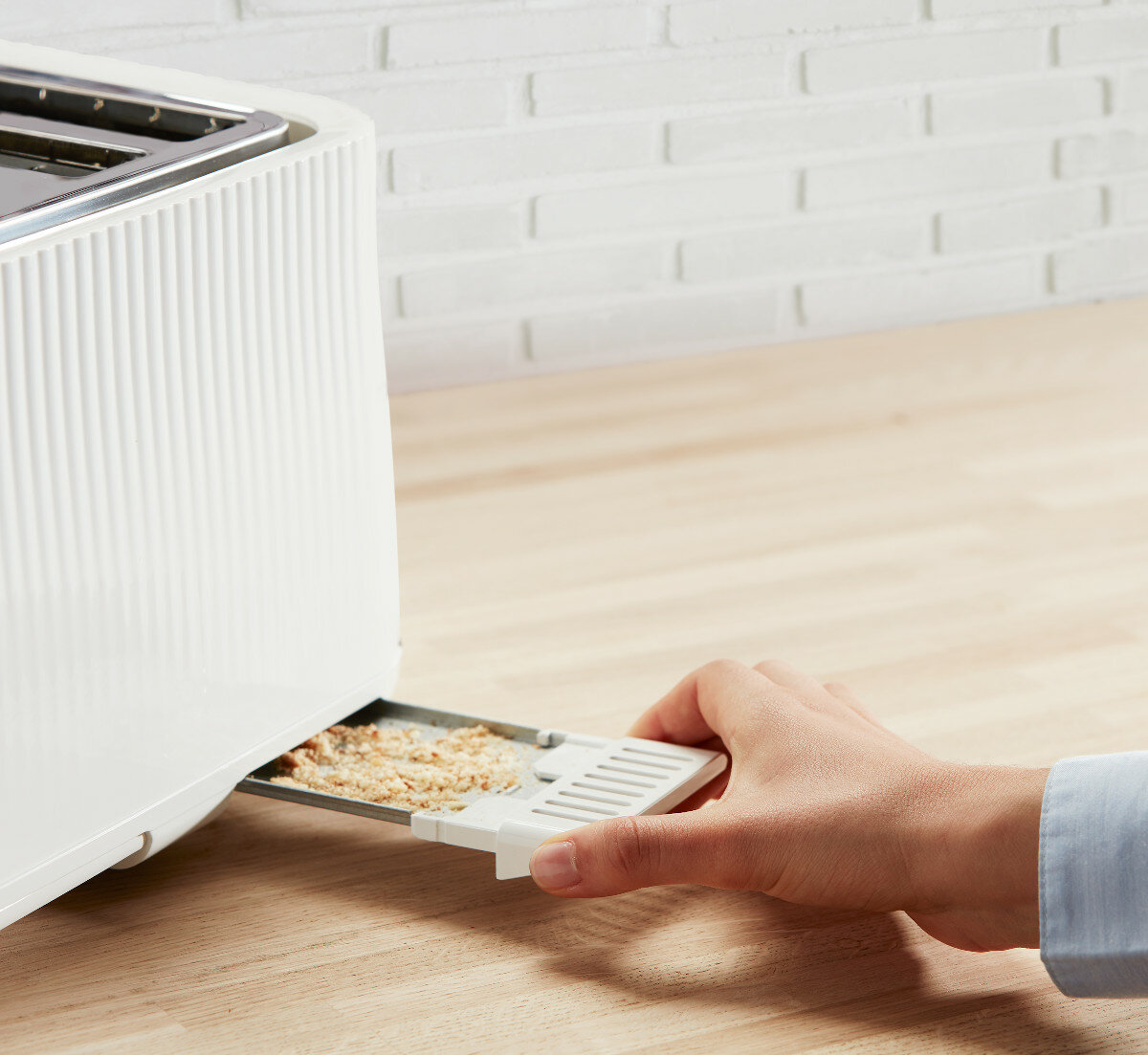 Ręka wyjmująca tackę na okruchy z białego tostera. Taca ma okruchy. Biała ceglana ściana i drewniana powierzchnia.