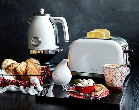 Scena kuchenna z białym czajnikiem, tosterem, chlebem, kawą i innymi produktami spożywczymi.