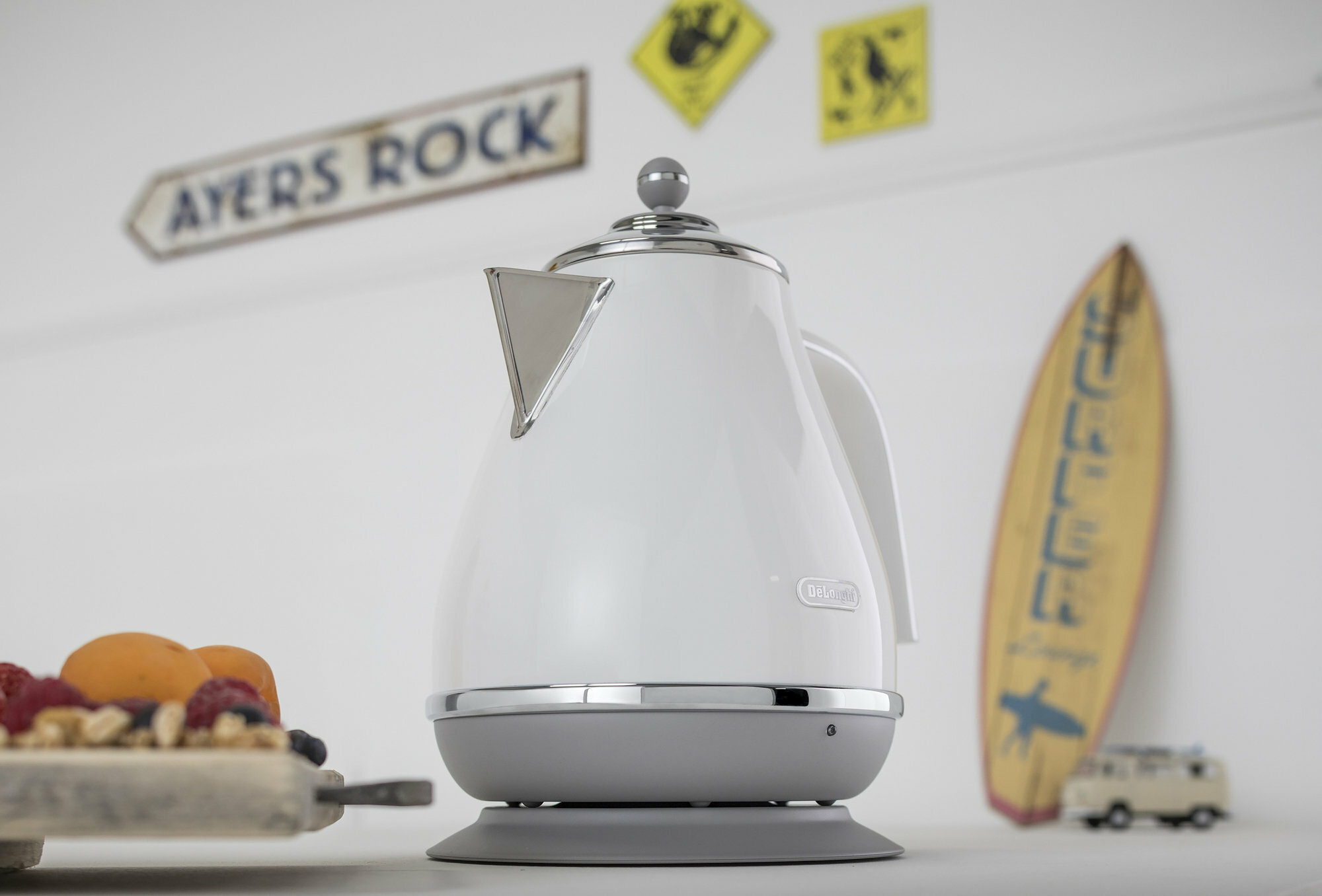 Biały czajnik elektryczny na szarej podstawie z deską surfingową i znakiem w tle.