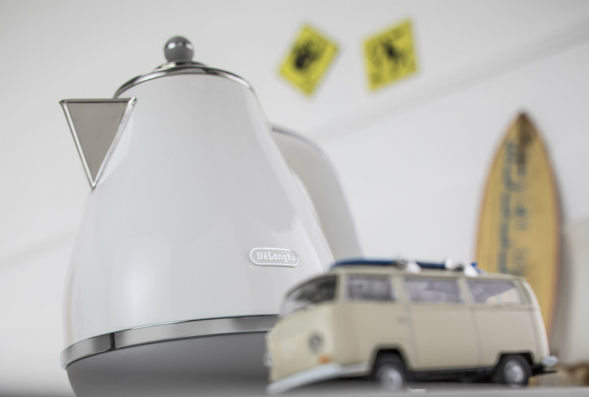 Biały czajnik DeLonghi ze srebrnym obramowaniem i zabawką van, z dwoma żółtymi znakami w tle.