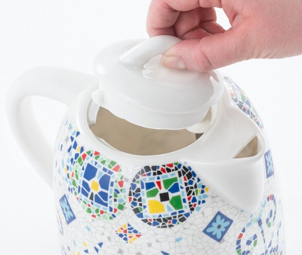 Ręka otwiera pokrywkę białego i kolorowego czajnika z wzorem mozaiki.
