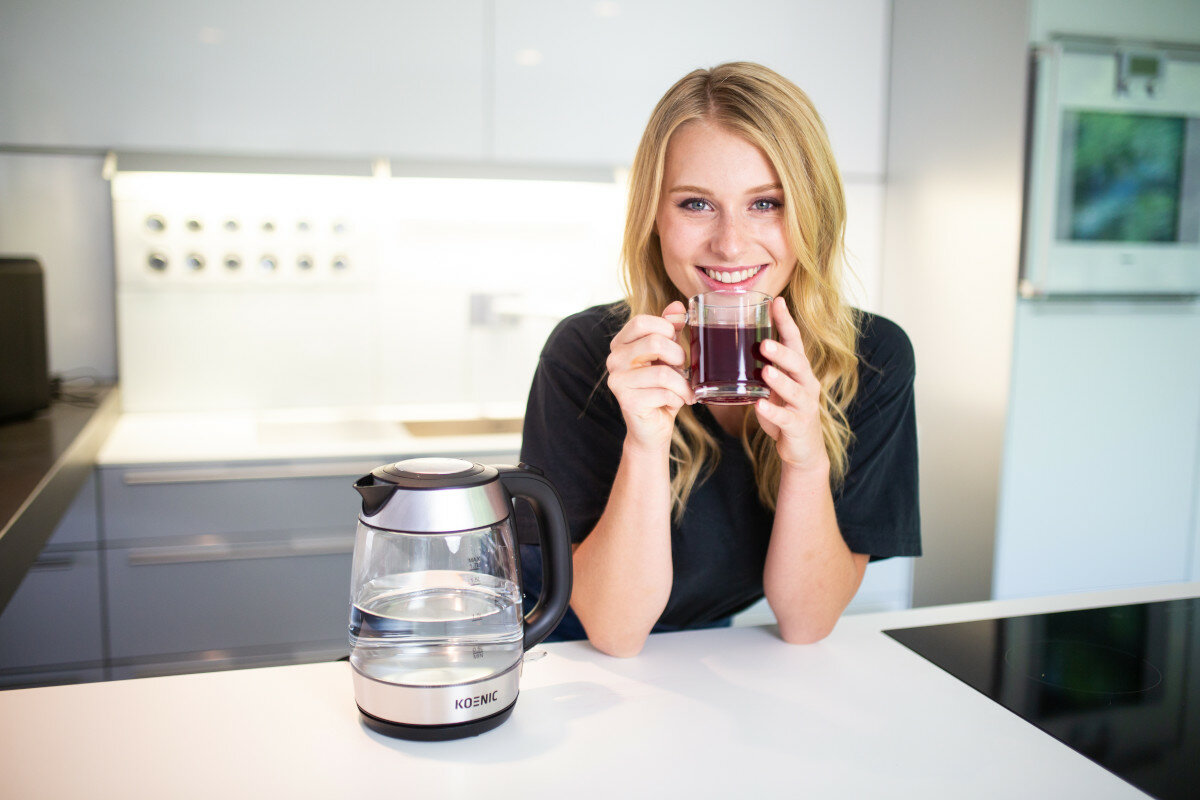 Kobieta trzymająca szklankę z czerwonym płynem i uśmiechająca się, z przezroczystym czajnikiem elektrycznym na białym blacie.