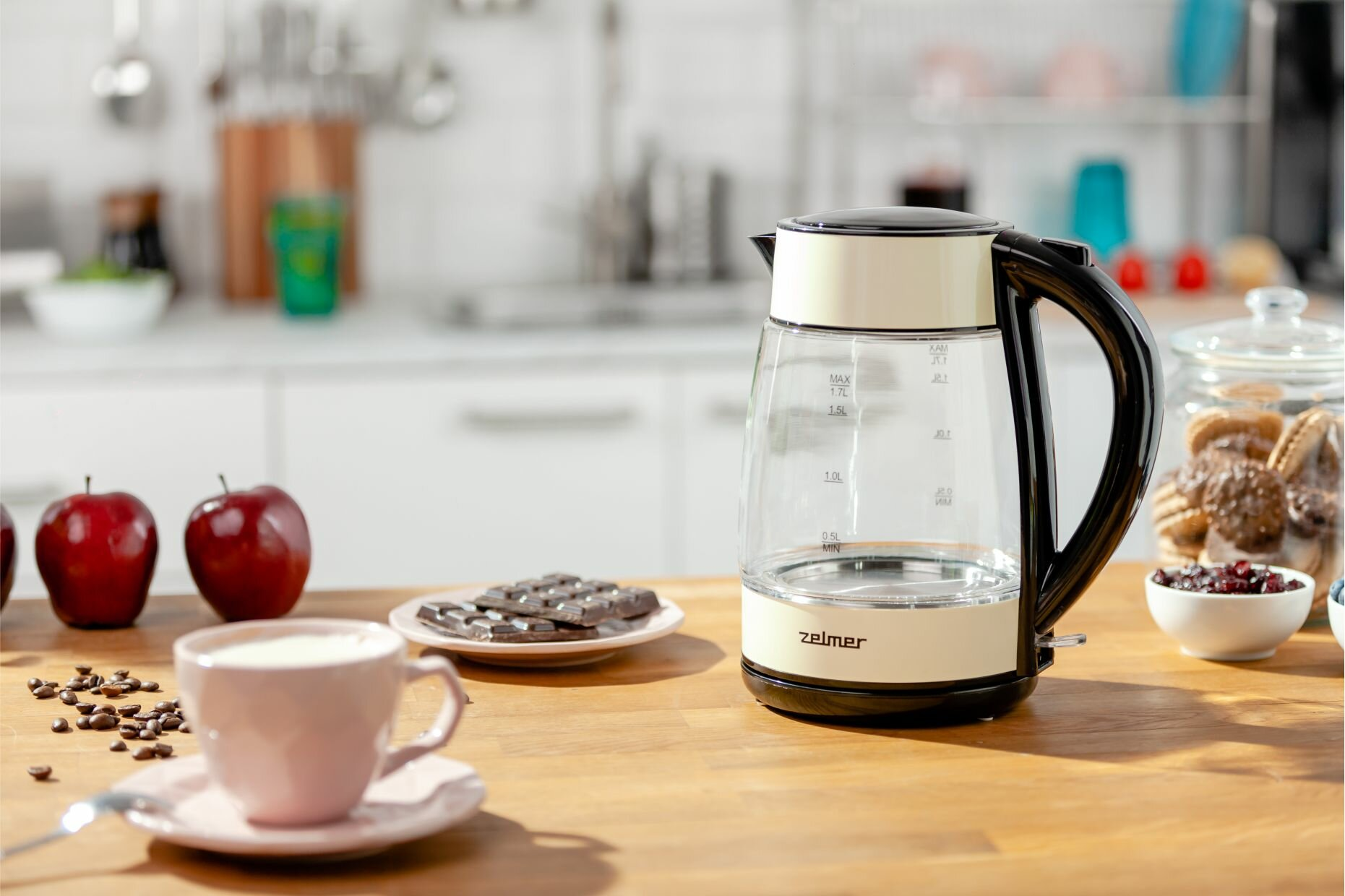 Szklany czajnik elektryczny i filiżanka kawy na drewnianym stole z jedzeniem i dekoracjami.