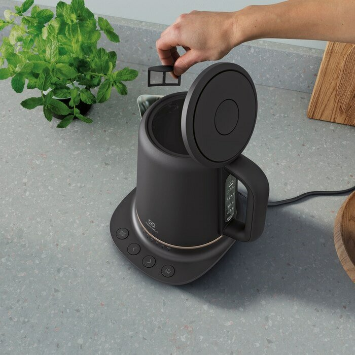 Czarny czajnik z otwartą pokrywą, dłoń dodająca filtr, blat kuchenny z roślinami.