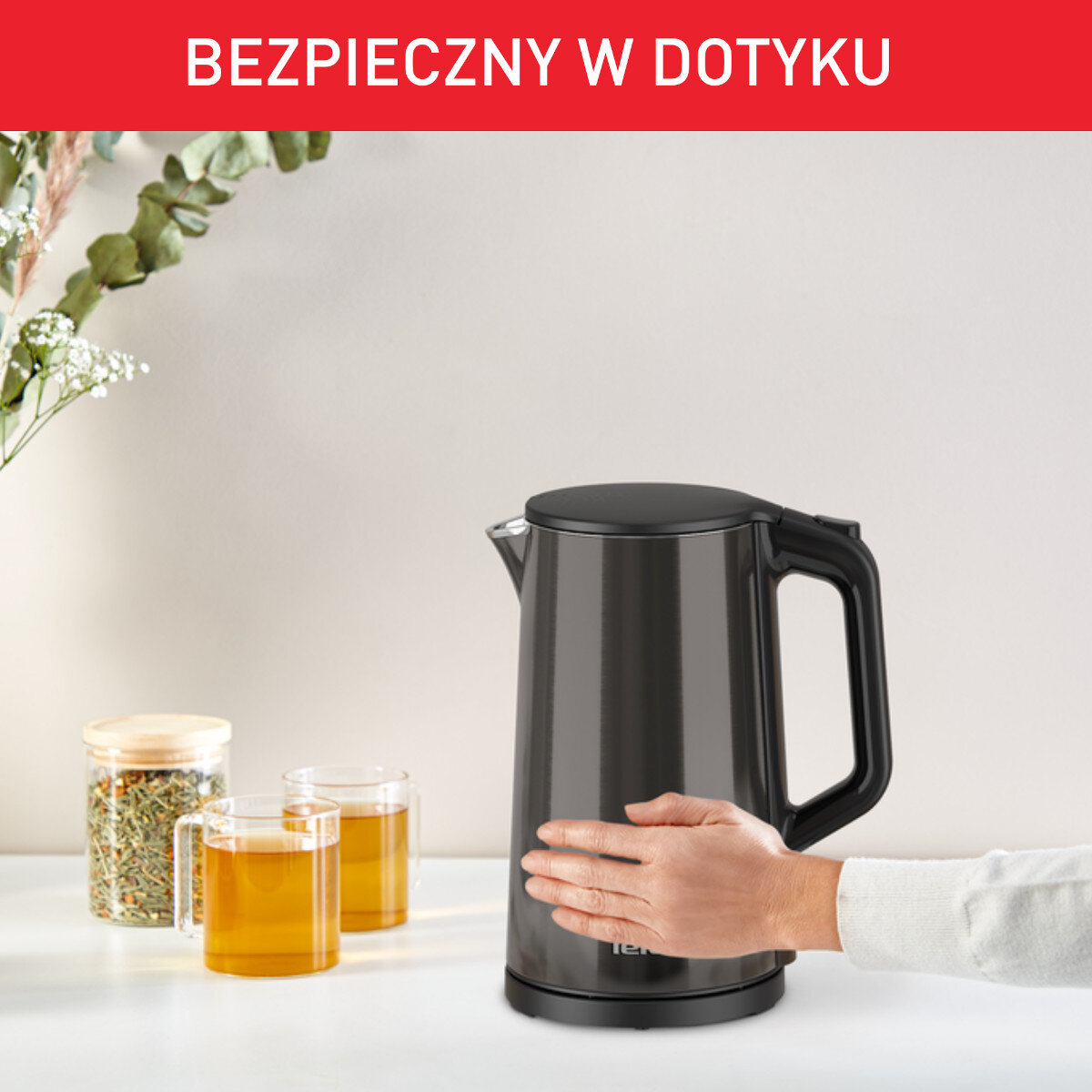 Czarny czajnik elektryczny z ręką na nim, obok dwóch szklanek herbaty.