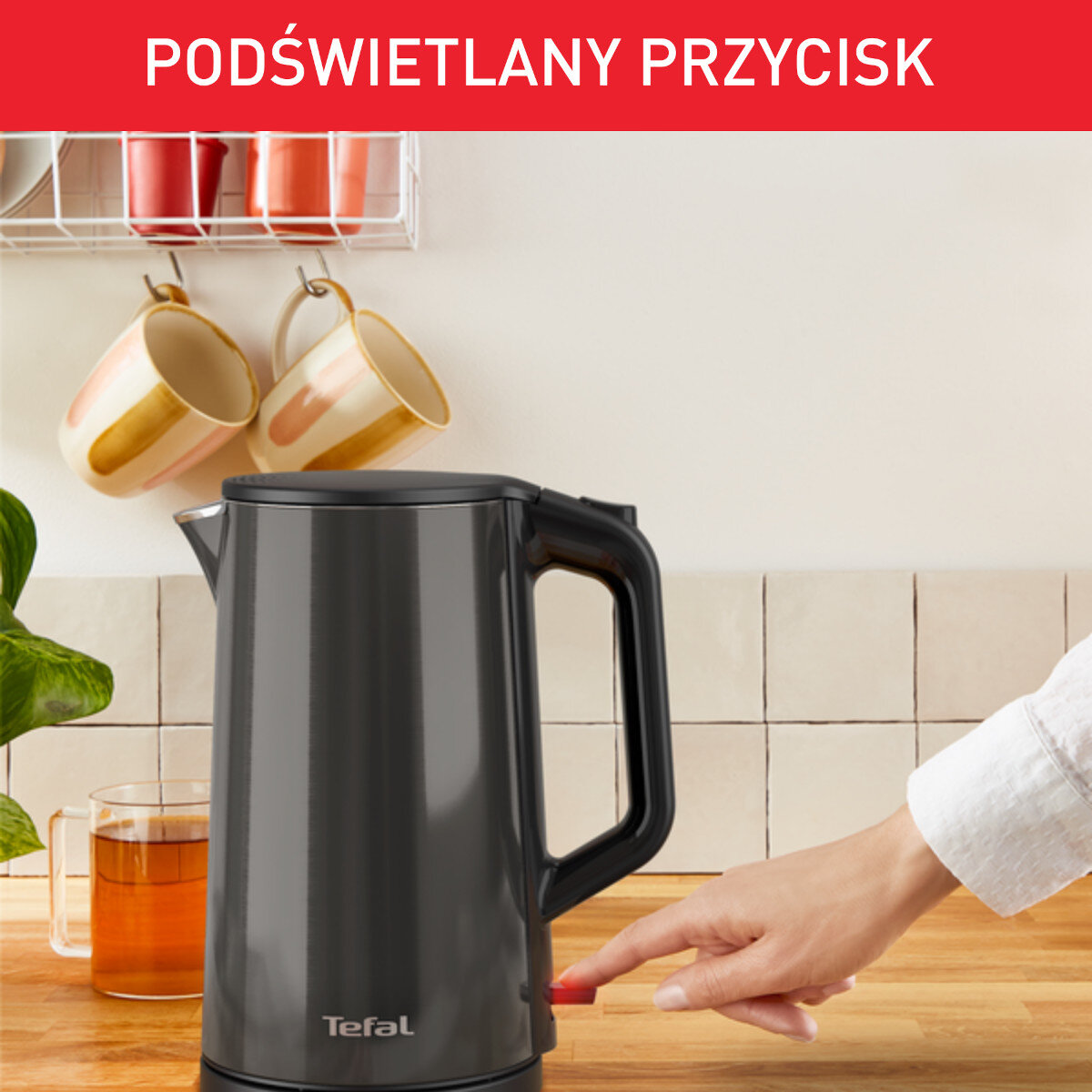 Czarny czajnik Tefal z podświetlanym czerwonym przyciskiem, na drewnianej powierzchni z kubkami.