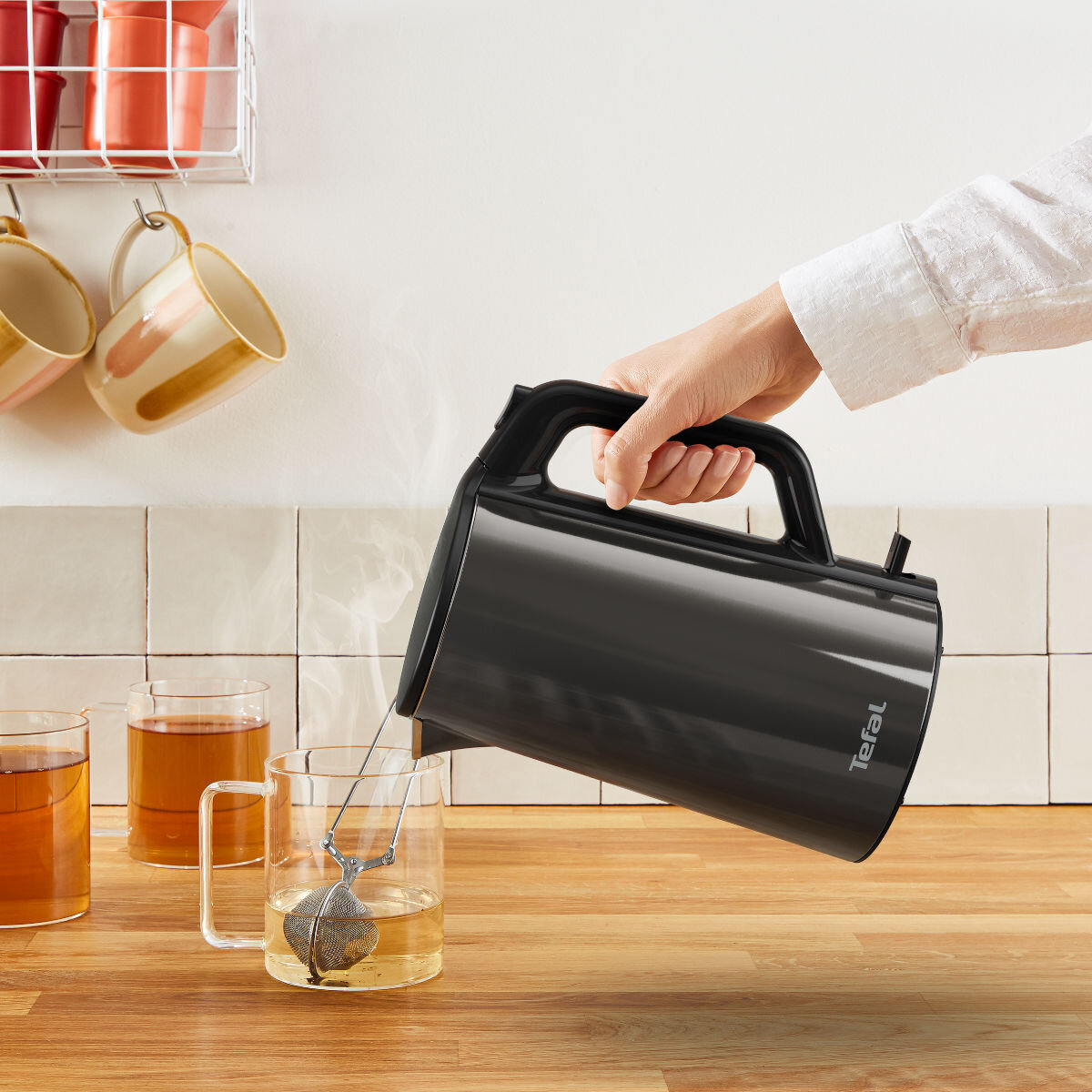 Osoba nalewa wodę z czarnego czajnika do szklanki z zaparzaczem do herbaty.
