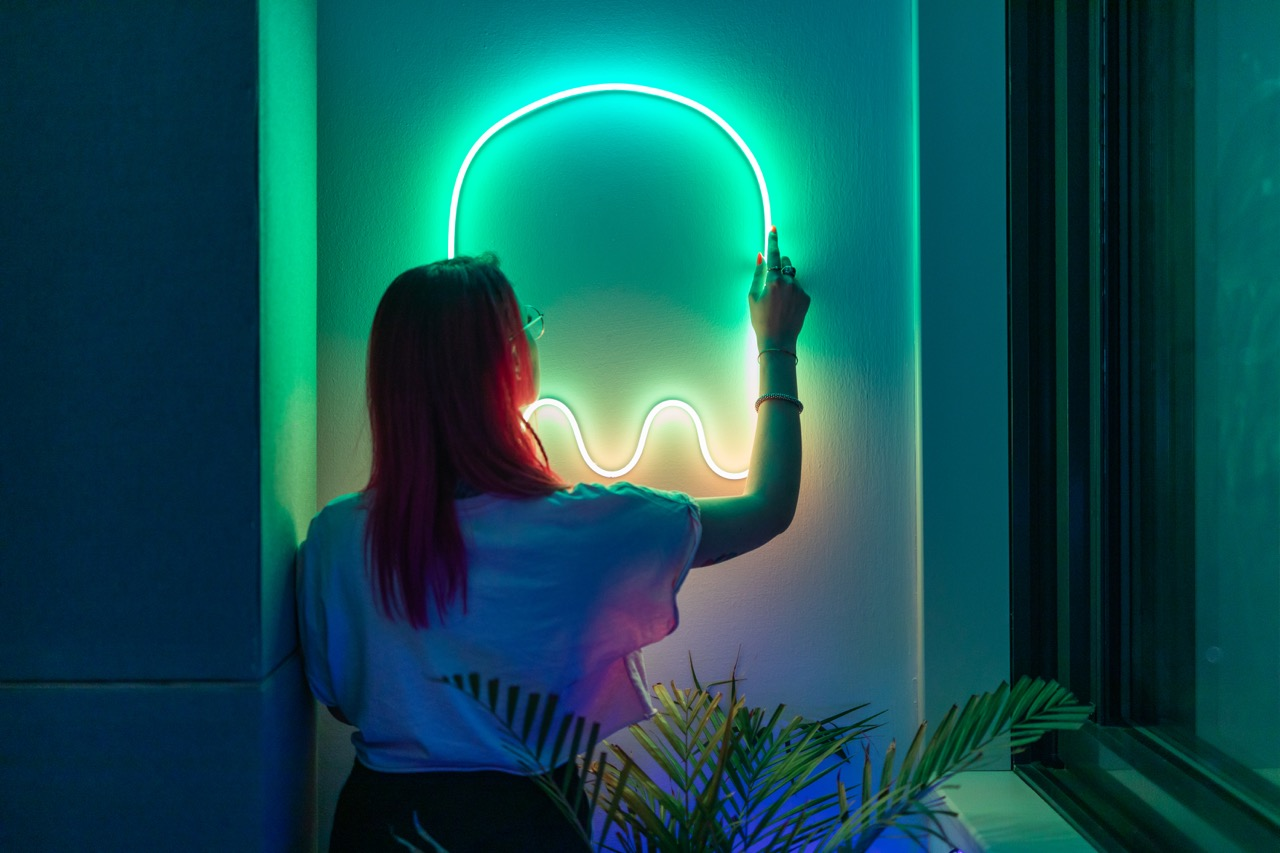 Donna che tocca un'insegna al neon a forma di fantasma. Capelli rosa, camicia bianca, sfondo verde e blu.