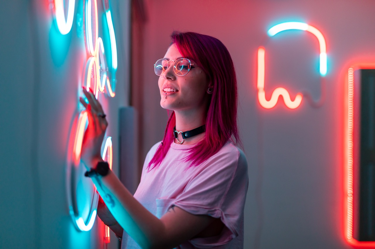 Una donna con i capelli rosa tocca un muro con luci al neon. La stanza è scarsamente illuminata.