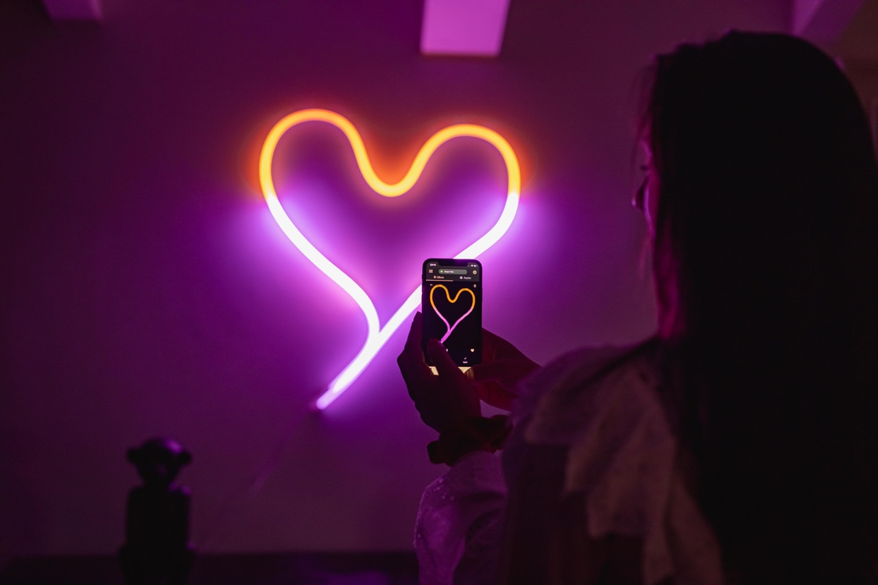 Una persona che tiene un telefono che mostra un contorno di cuore, con un cuore al neon sulla parete dietro di loro.