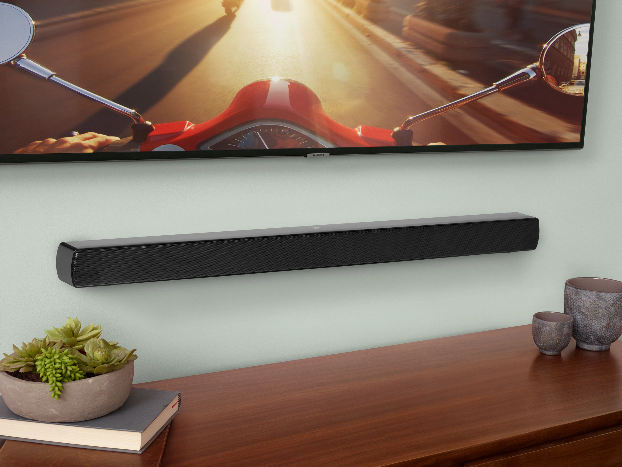 Schwarze Soundbar an einer mintfarbenen Wand, unter einem Fernseher mit Roller. Holzschrank, Pflanze und Deko.