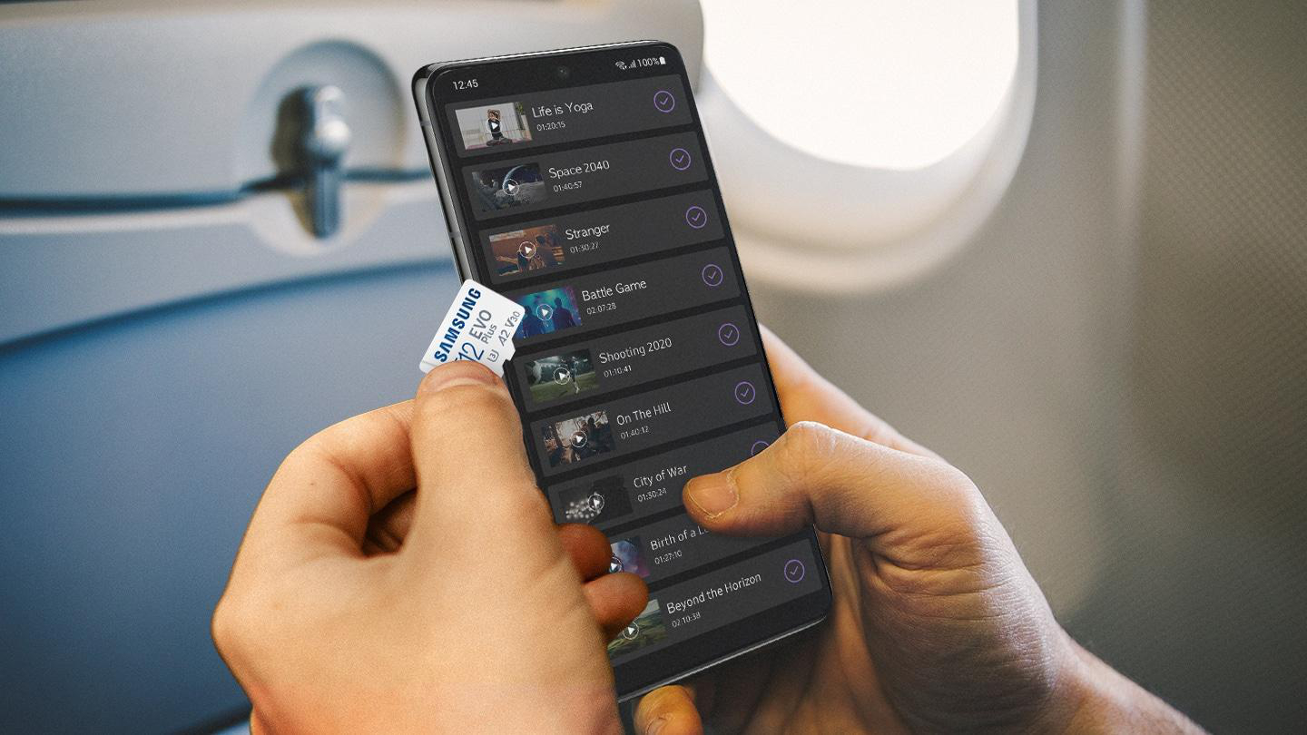 Eine Person hält ein Telefon mit Videotiteln und eine Samsung-Speicherkarte. Der Hintergrund zeigt ein Flugzeugfenster.