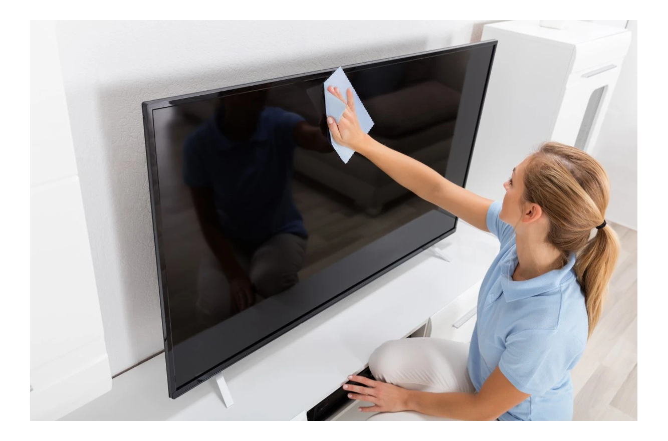 Une femme nettoyant un grand écran de télévision sur un meuble blanc. La télévision est éteinte.