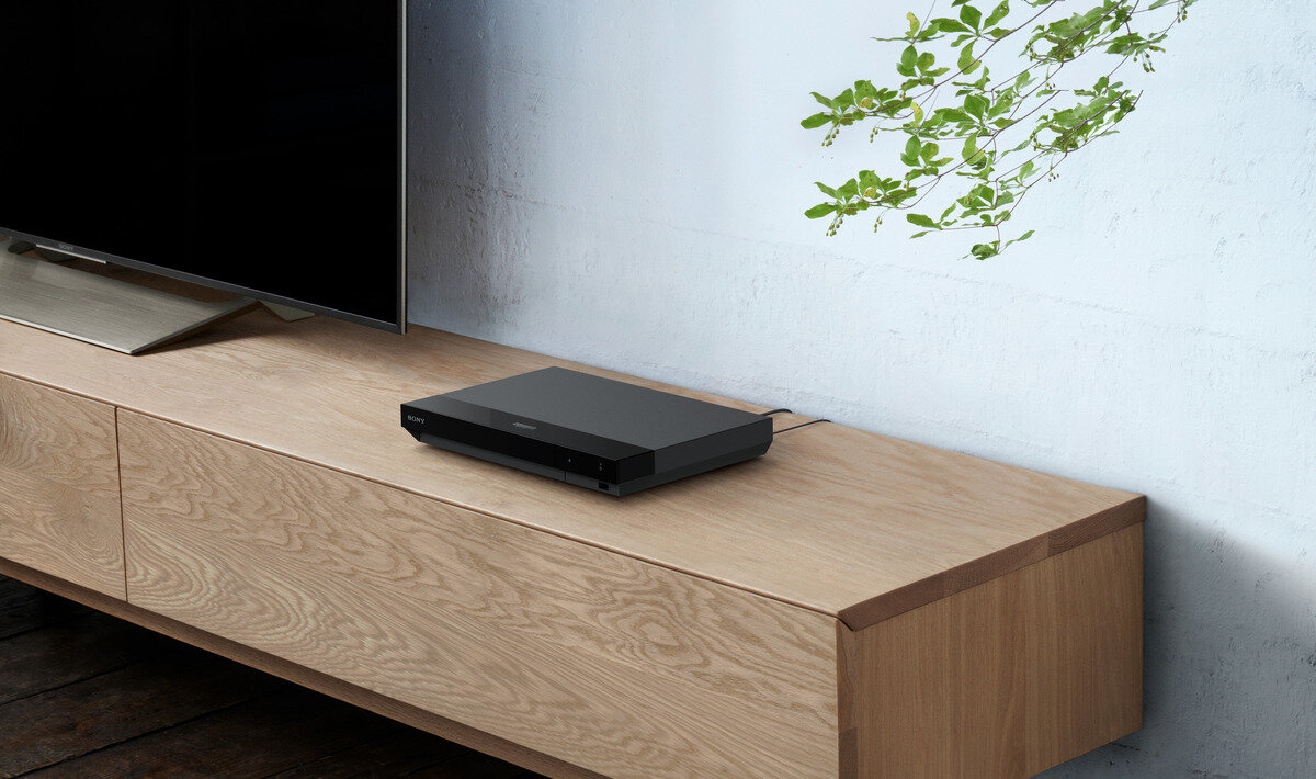 Czarny odtwarzacz Blu-ray Sony na drewnianej szafce, z telewizorem w tle i rośliną.