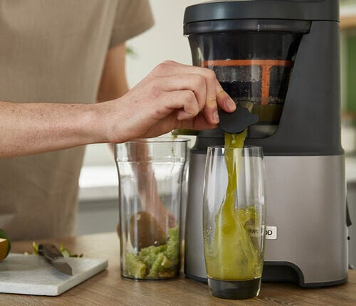 Osoba wlewa zielony sok z wyciskarki do szklanki. Widoczna jest kolejna szklanka i owoce.
