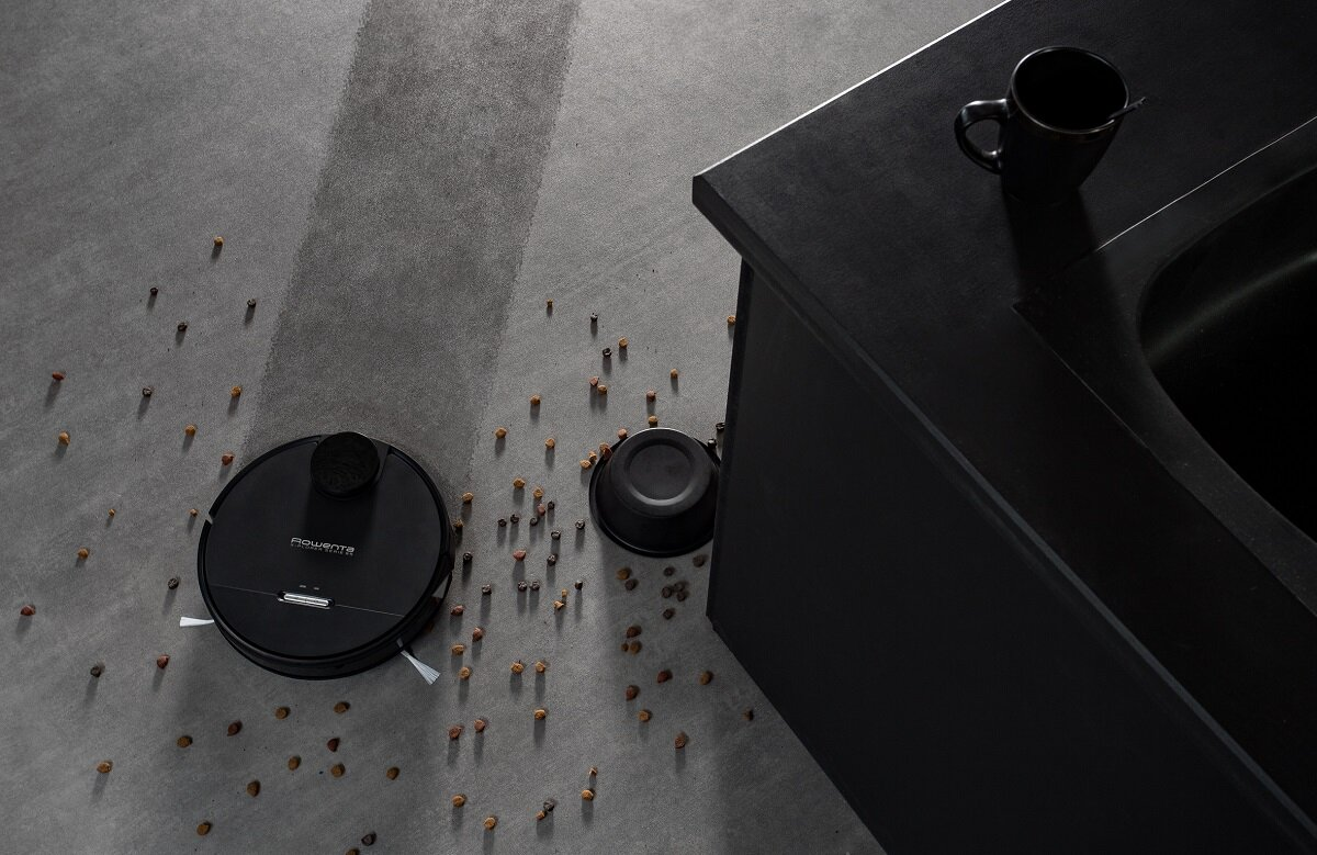 Odkurzacz robot na szarej podłodze z rozrzuconymi śmieciami w pobliżu czarnej szafki.