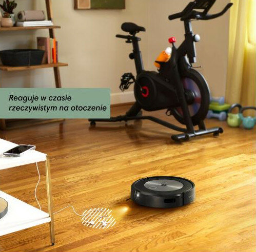 Czarny odkurzacz robot na drewnianej podłodze. W tle rower stacjonarny.