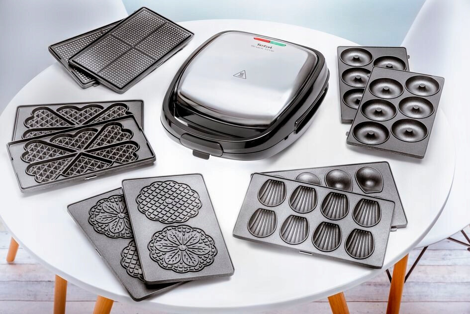 Gofrownica z różnymi płytami, w tym kształty gofrów, pączków i madeleines. Umieszczona na białej powierzchni.