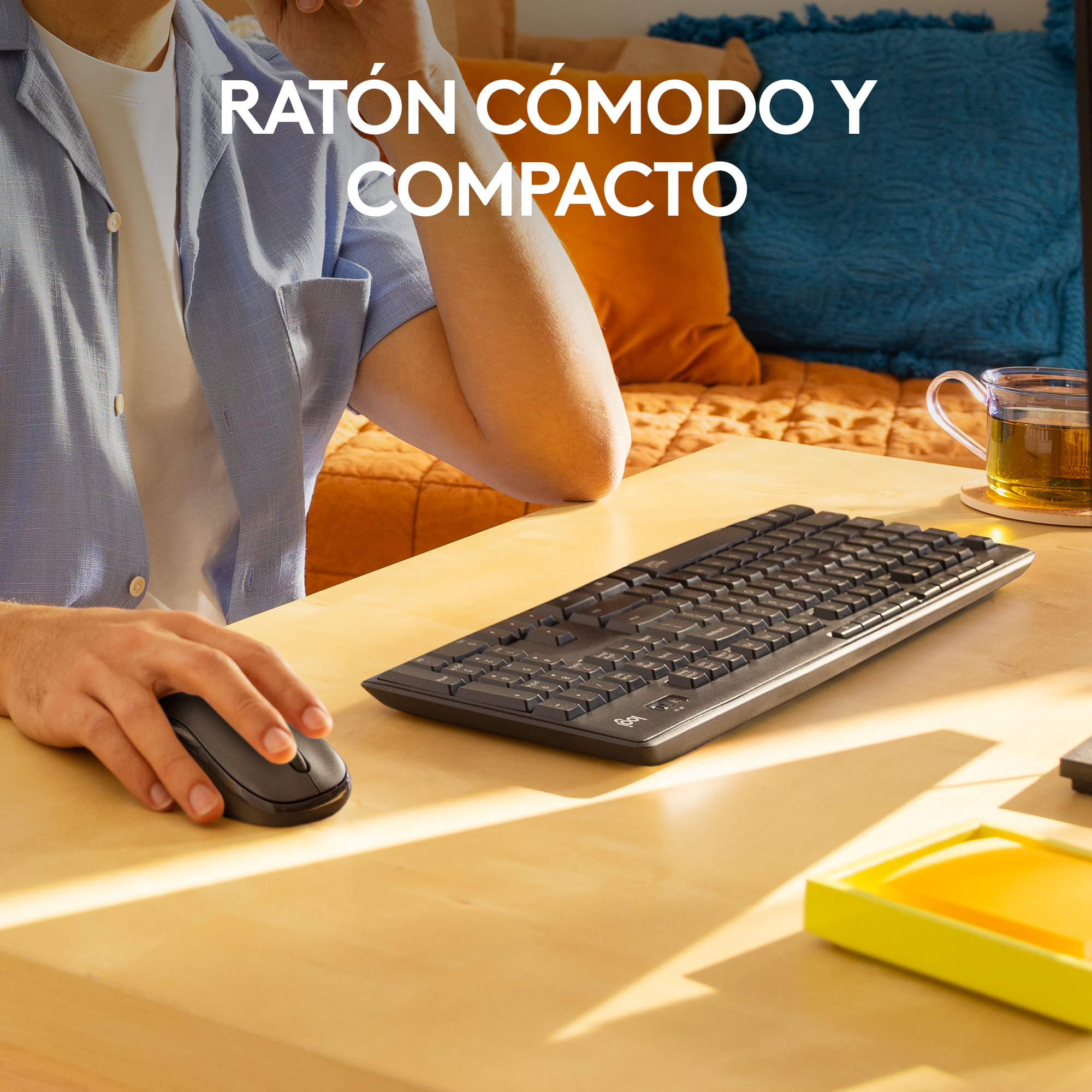 Una persona usa un ratón y teclado negros en un escritorio de madera. Almohadas azules y naranjas.