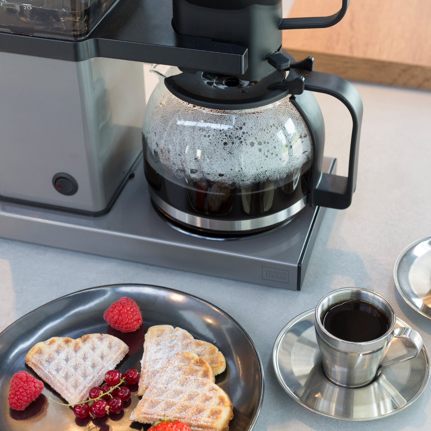 Een koffiezetapparaat met een glazen kan. Wafels, bessen en een kopje koffie staan op tafel.