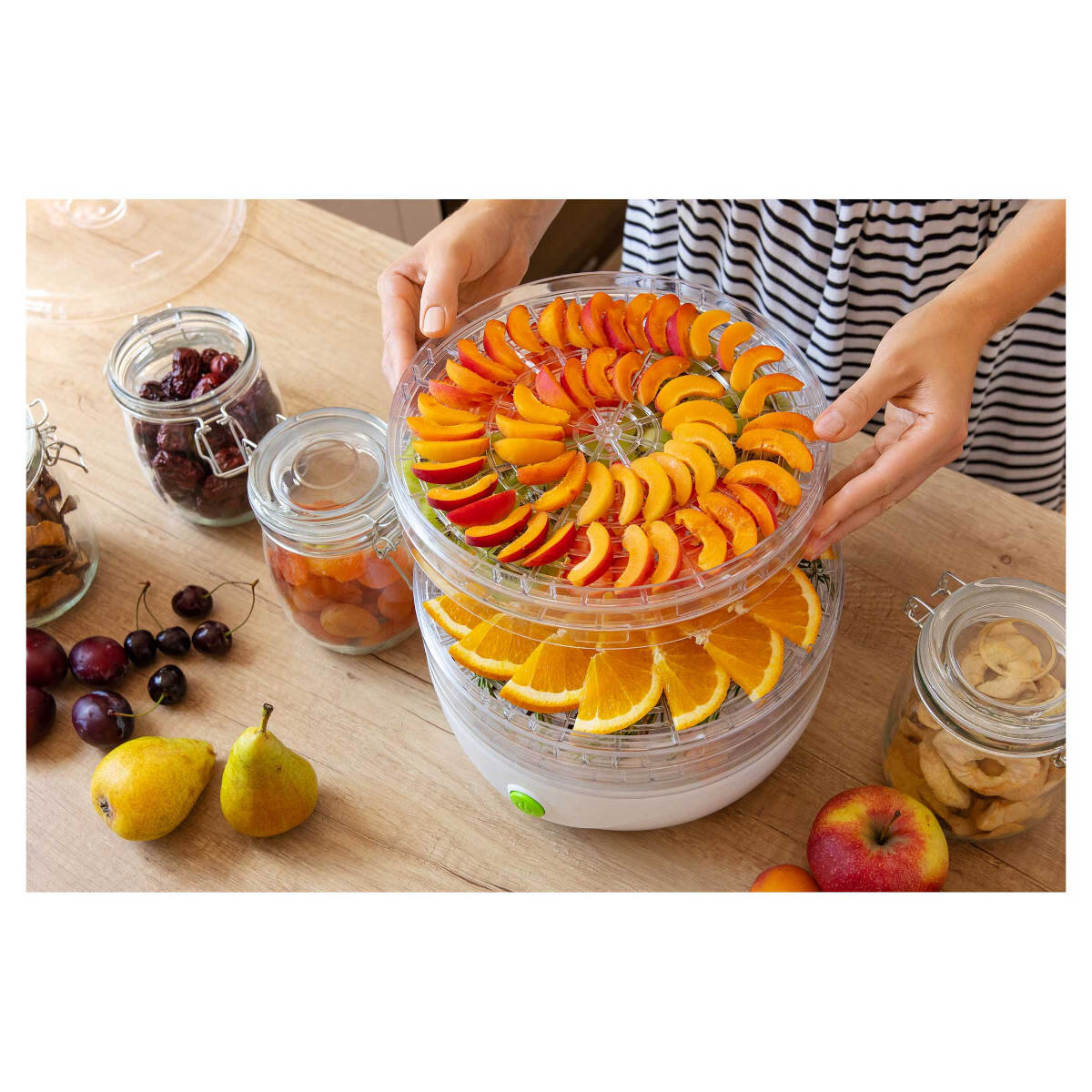 Osoba trzyma tacę z suszarki z pokrojonymi morelami i pomarańczami. Słoiki z suszonymi owocami i świeżymi owocami na stole.