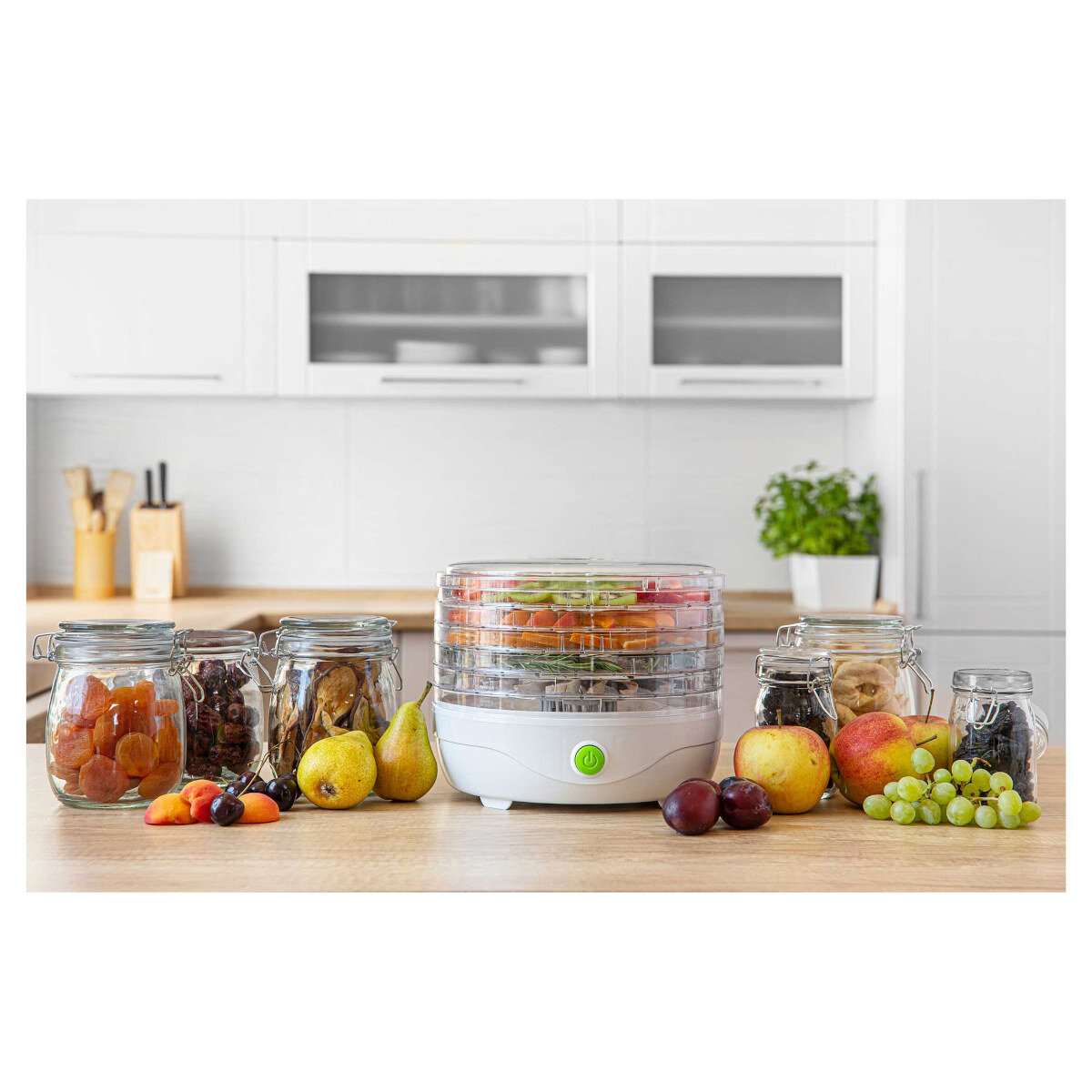 Suszarka do żywności z owocami i słoikami na drewnianym stole. Tło: białe szafki kuchenne i roślina. Owoce: jabłka, winogrona, śliwki, gruszki.