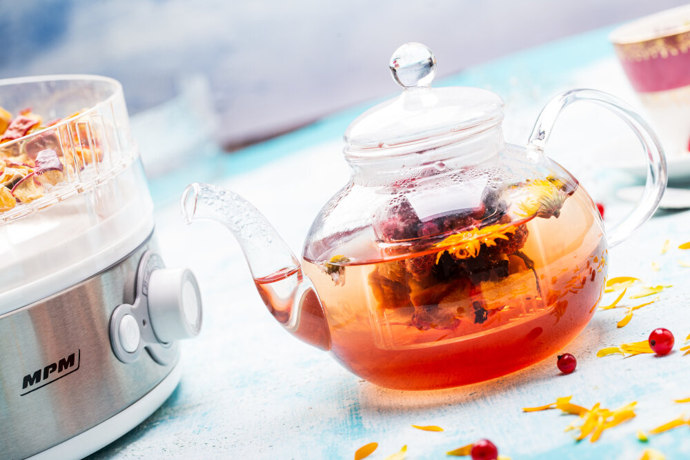 Szklany czajniczek z herbatą i kwiatami, obok zaparzacza do herbaty. Niebieskie tło.