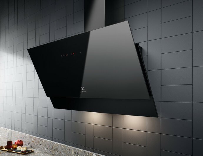 Czarny, nowoczesny okap zamontowany na szarej, kafelkowanej ścianie, z blatem i tacą z jedzeniem.