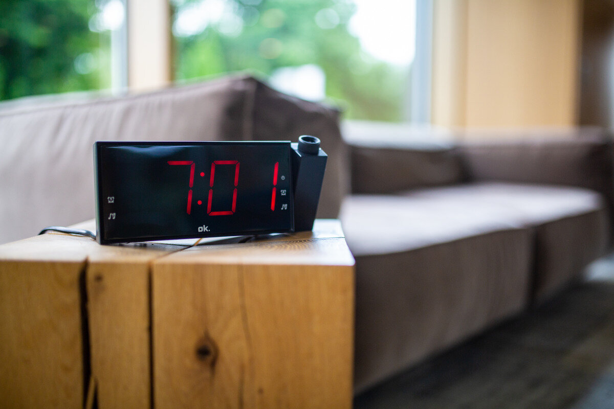 Budzik cyfrowy wyświetlający godzinę 7:01, stojący na drewnianej szafce nocnej obok brązowej sofy.