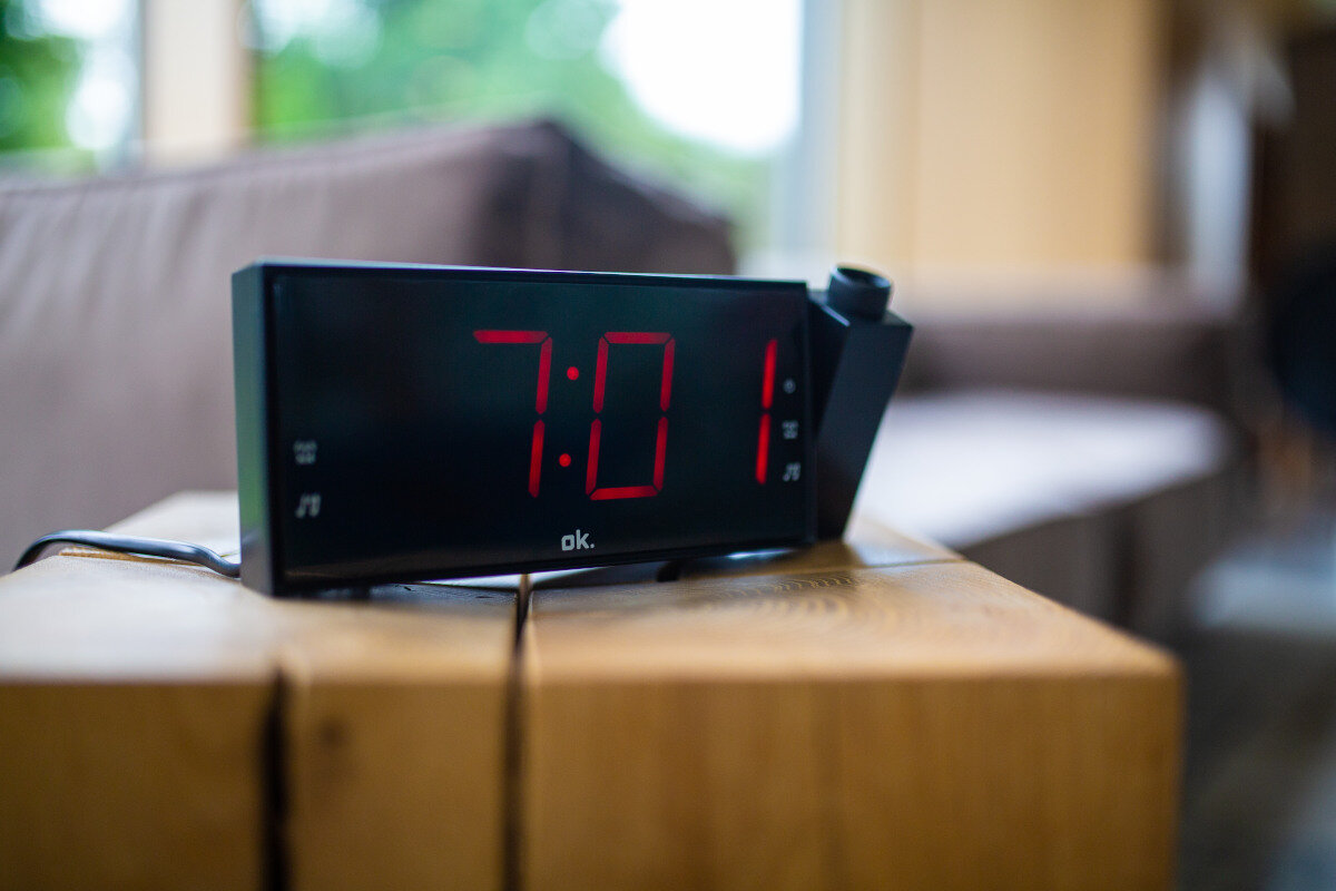 Czarny cyfrowy budzik pokazuje 7:01 na drewnianym stoliku, rozmyte tło.