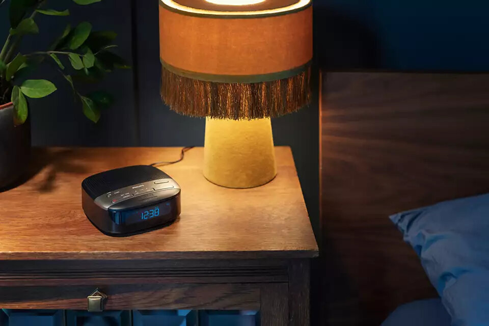 Cyfrowy budzik na drewnianej szafce nocnej. Nad nim zapalona lampa.