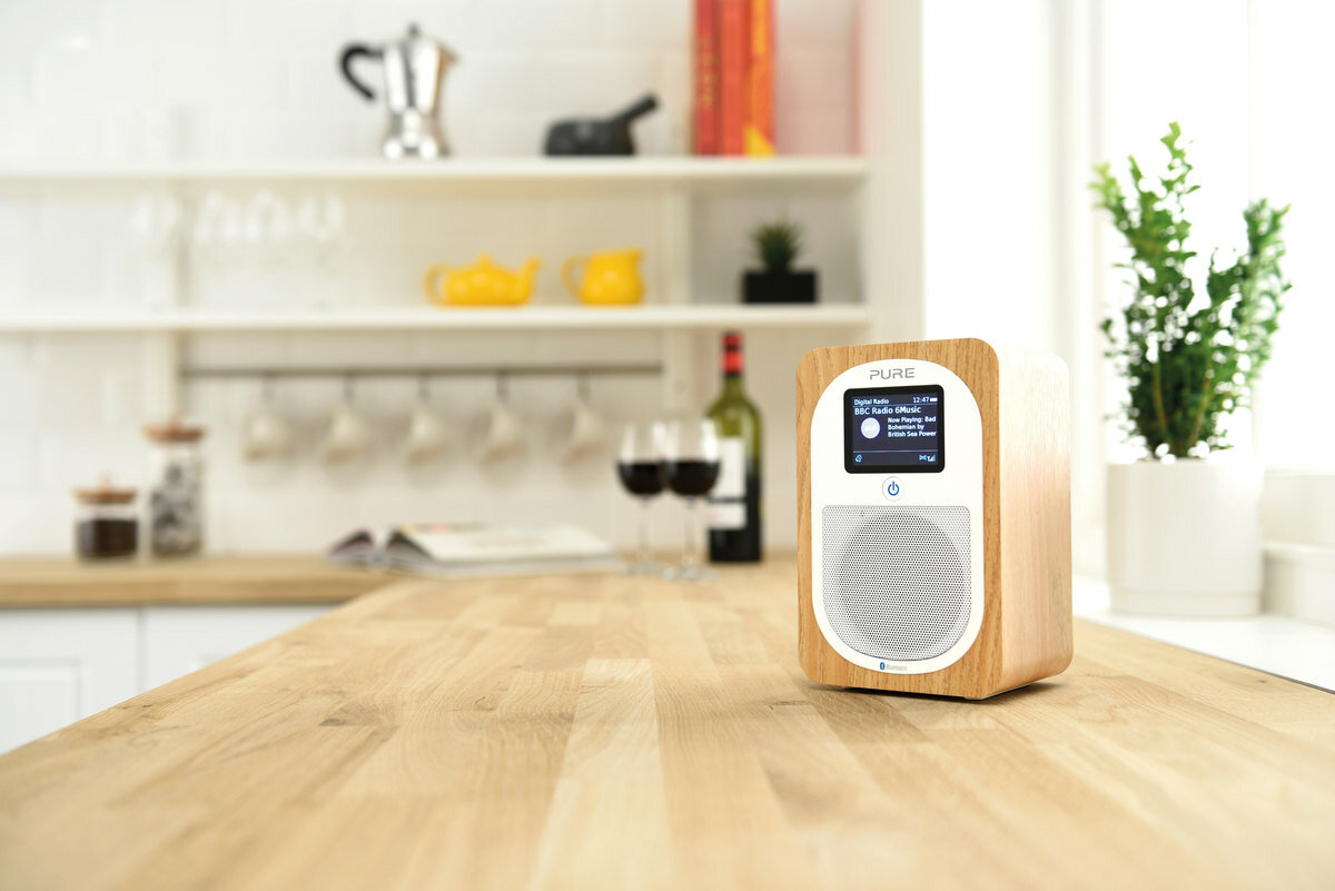 Drewniane radio z małym ekranem, na drewnianym blacie. W tle scena kuchenna.