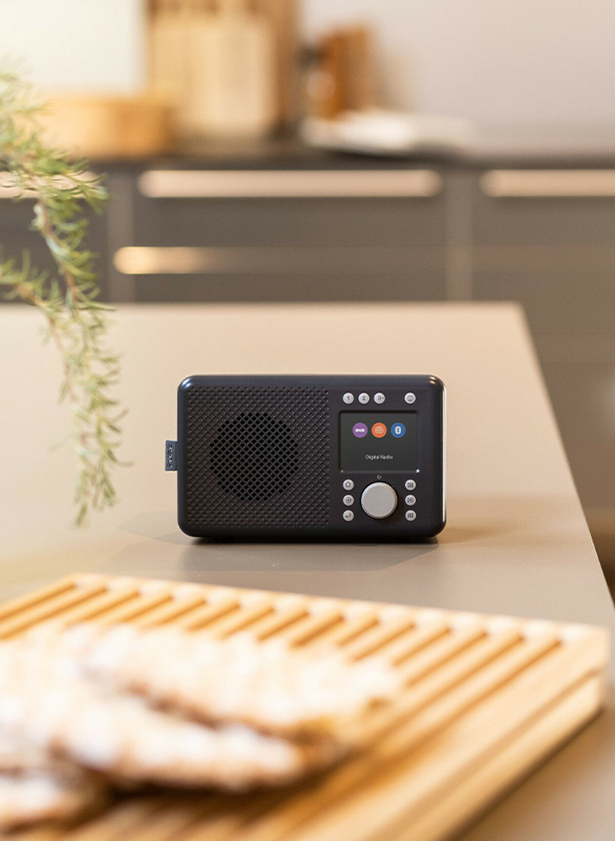 Czarny radio cyfrowe na jasnobrązowym stole, z jedzeniem i zielenią w tle.