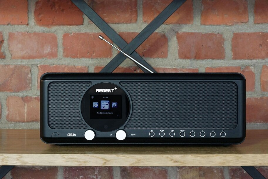 Czarny radio z ekranem, przyciskami i anteną na drewnianej półce.